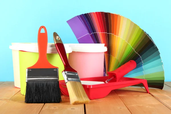 Paint pots, paintbrushes and coloured swatches on wooden table on blue background — Stock Photo, Image