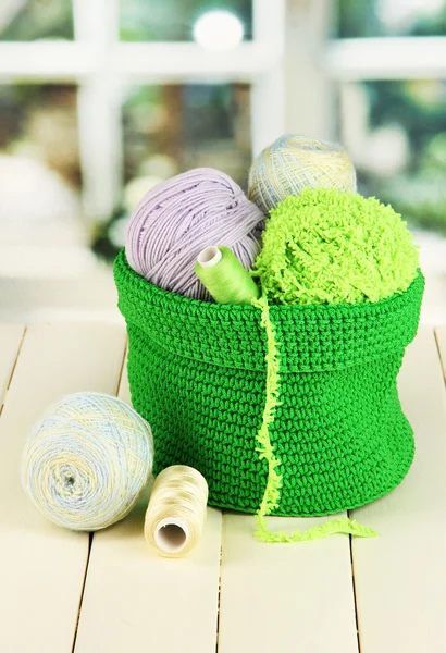 Красочная пряжа для вязания в зеленой корзине на деревянном столе на фоне окна — стоковое фото