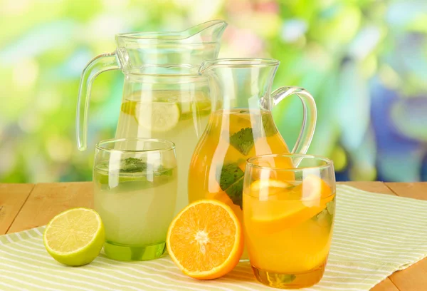 Orange and lemon lemonade in pitchers and glasses on wooden table on natural background — Stock Photo, Image