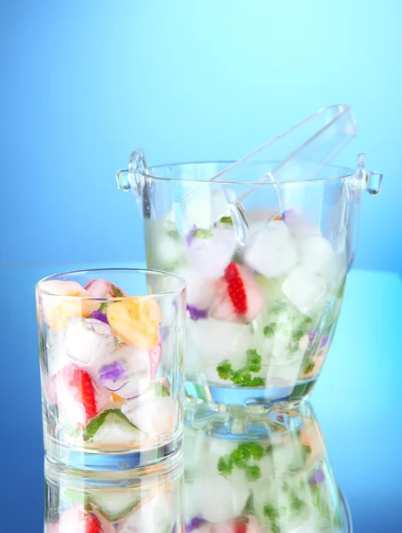 Ледяные кубики с цветами, фруктами и травами в стеклянном и стеклянном ведре, на синем фоне — стоковое фото
