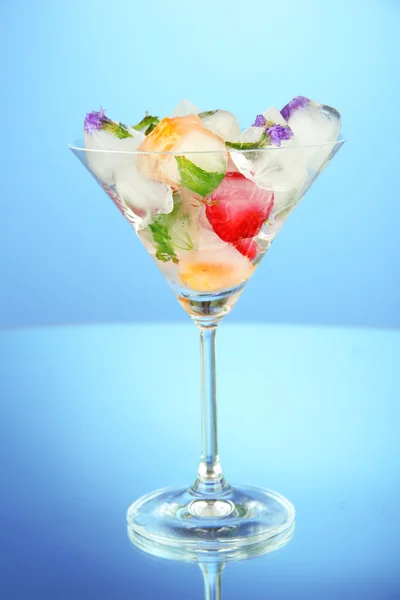 Eiswürfel mit Blumen, Kräutern und Früchten im Inneren in Martini-Glas, auf blauem Hintergrund — Stockfoto