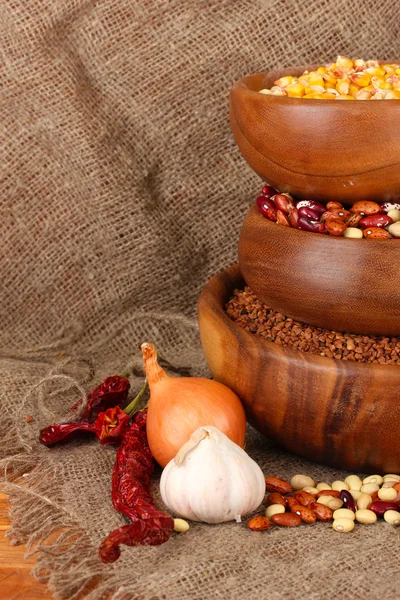Сира кукурудза, гречка і квасоля в дерев'яних мисках на столі на тлі мішковини — стокове фото