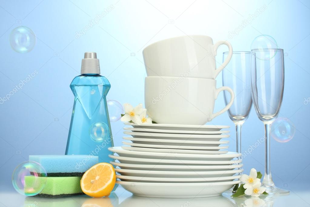empty clean plates, glasses and cups with dishwashing liquid, sponges and lemon on blue background