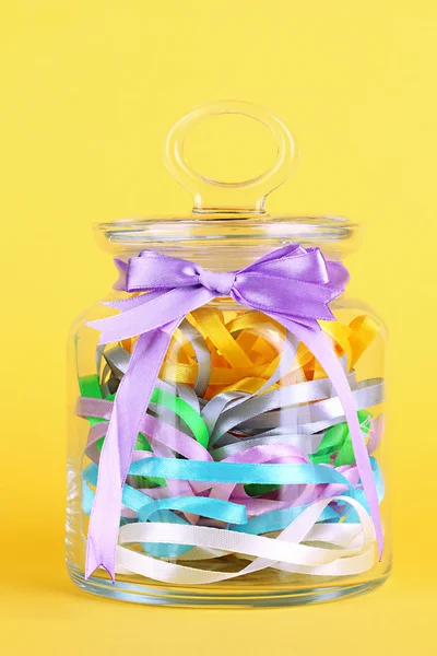 Glass jar containing various colored ribbons on yellow background — Stock Photo, Image