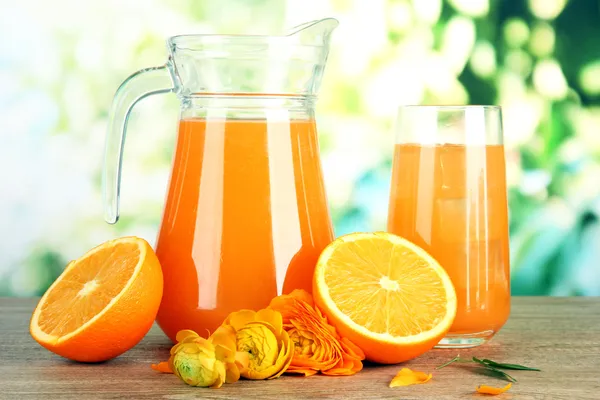 Glass and pitcher of orange juice on wooden table, on green background — Stock Photo, Image