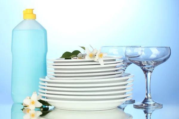 Pratos limpos vazios e copos com líquido de lavagem de louça no fundo azul — Fotografia de Stock