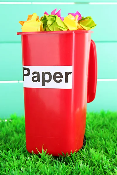 Recycling bin with papers on grass on light blue background — Stock Photo, Image