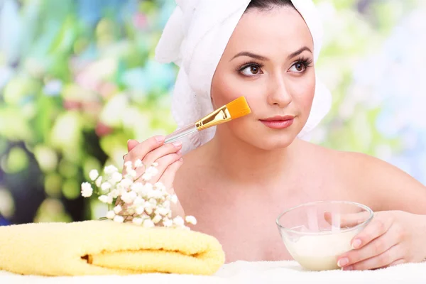 Beautiful young woman with cream for face mask towel on her head on bright background — Stock Photo, Image