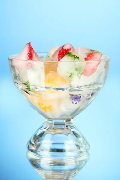 Cubetti di ghiaccio con fiori, pezzi di frutta ed erbe aromatiche in vaso di vetro, su sfondo blu — Foto Stock