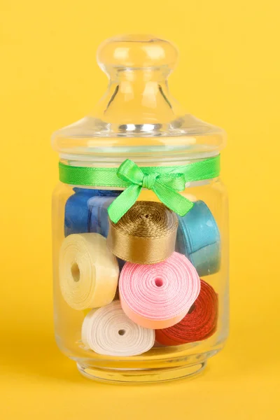 Glass jar containing various colored ribbons on yellow background — Stock Photo, Image