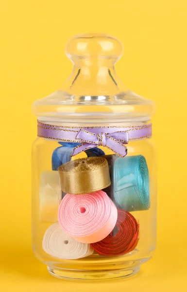 Glass jar containing various colored ribbons on yellow background — Stock Photo, Image