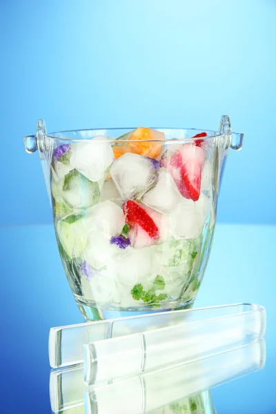 Ledové kostky s květy, kousky ovoce a bylin v kbelíku skla, na modrém pozadí — Stock fotografie