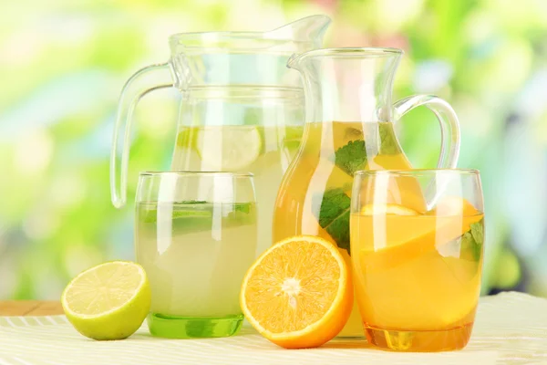 Orange and lemon lemonade in pitchers and glasses on wooden table on natural background — Stock Photo, Image