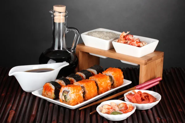 Delicioso sushi en el plato, palillos, salsa de soja, pescado y camarones en la estera de bambú sobre fondo gris —  Fotos de Stock
