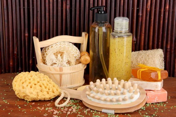 Limpador, escova e cosméticos para chuveiro na mesa de madeira sobre fundo de bambu — Fotografia de Stock