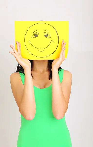 Young woman holding paper with happy smiley on gray background. — Stock Photo, Image