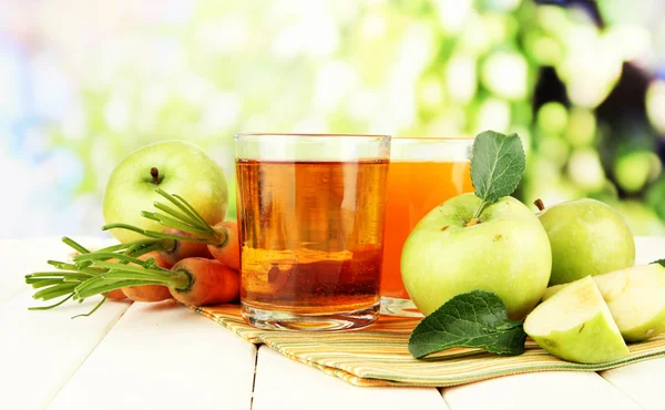 Glasses of juice, apples and carrots on white wooden table, on green background — Stock Photo, Image