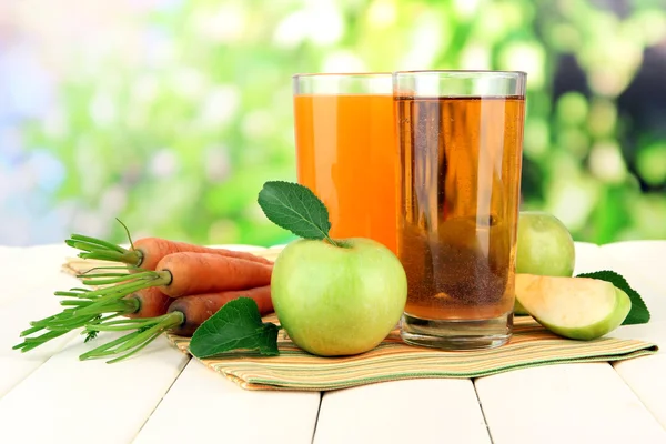 Glazen van SAP, appelen en wortelen op witte houten tafel, op groene achtergrond — Stockfoto