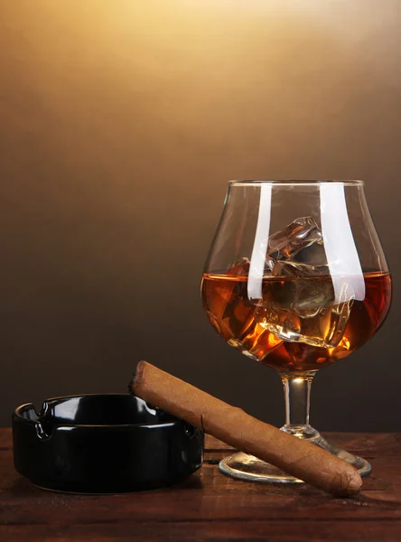 Brandy glass with ice and cigar on wooden table on brown background — Stock Photo, Image
