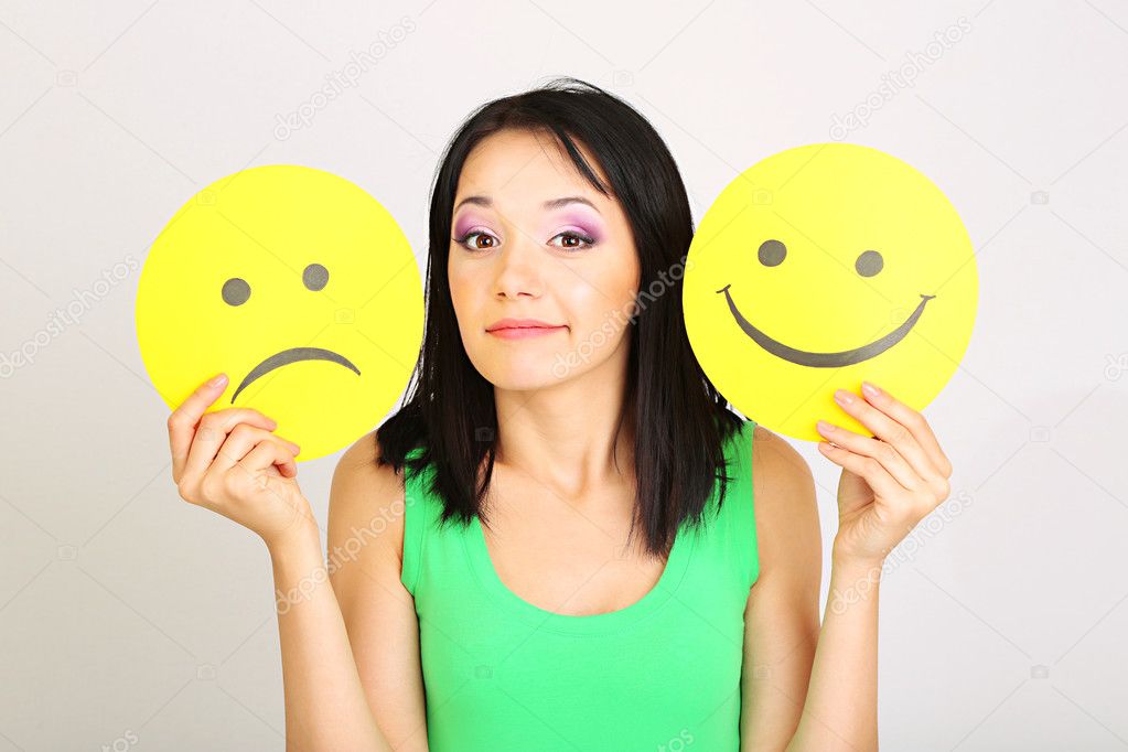 Young woman holding paper with sad and happy smiley on gray background