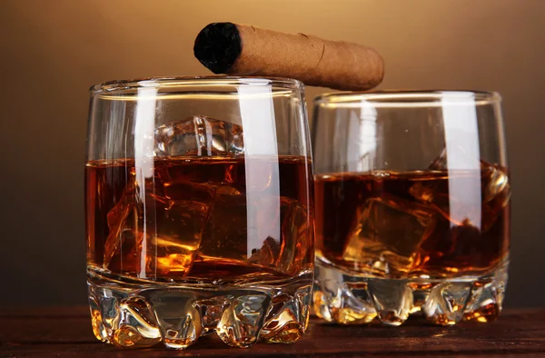 Brandy sklenice s ledem a doutník na dřevěný stůl na hnědé pozadí — Stock fotografie