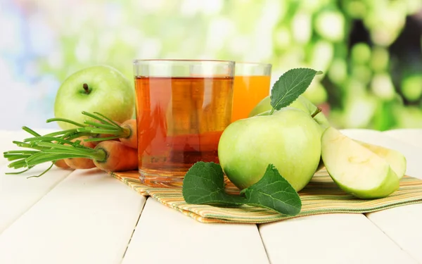 Saftgläser, Äpfel und Möhren auf weißem Holztisch, auf grünem Hintergrund — Stockfoto