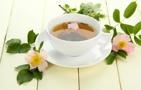 Φλιτζάνι τσάι βοτάνων με ισχίου ροδαλά λουλούδια στο λευκό ξύλινο τραπέζι — Φωτογραφία Αρχείου