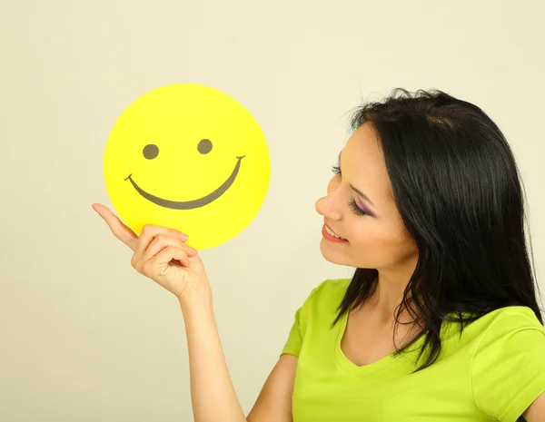 Ung kvinna med papper med glad smiley på grå bakgrund. — Stockfoto