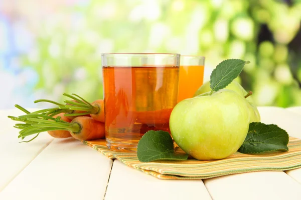 Glazen van SAP, appelen en wortelen op witte houten tafel, op groene achtergrond — Stockfoto