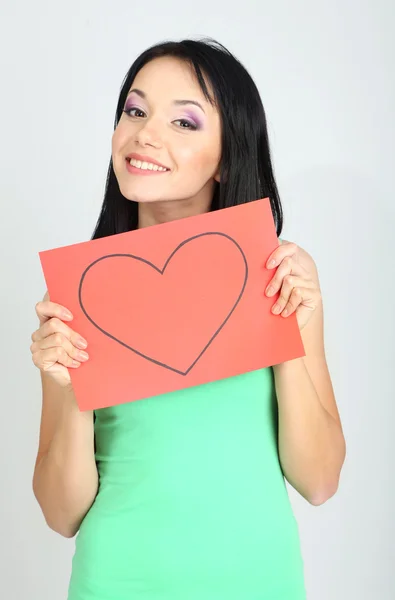 Young woman holding red painted heart on grey background — Stock fotografie