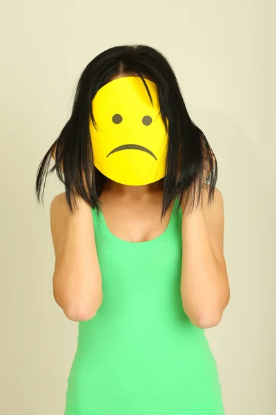 Young woman holding paper with sad smiley on gray background — Stock Photo, Image