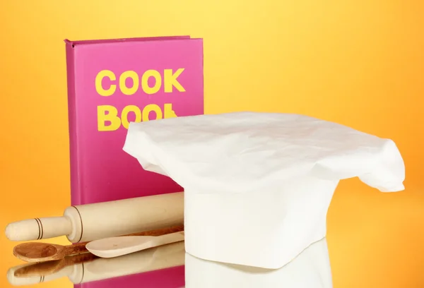 Chef's hat with battledore and cook book on orange background — Stock Photo, Image