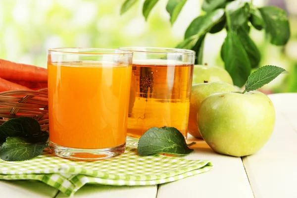 Glasses of juice, apples and carrots on white wooden table, on green background