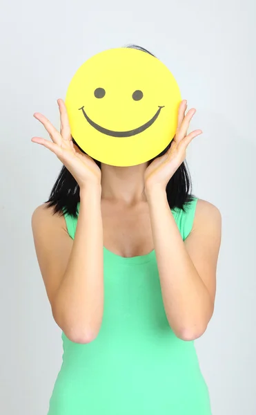 Junge Frau hält Papier mit fröhlichem Smiley auf grauem Hintergrund. — Stockfoto