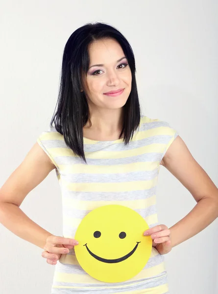 Young woman holding paper with happy smiley on gray background. — Stock Photo, Image