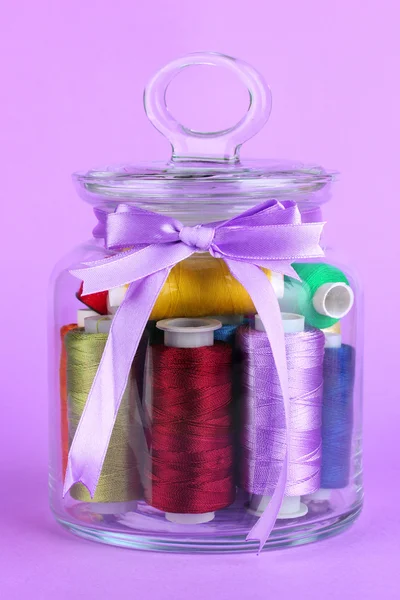 Glass jar containing various colored thread on lilac background — Stock Photo, Image