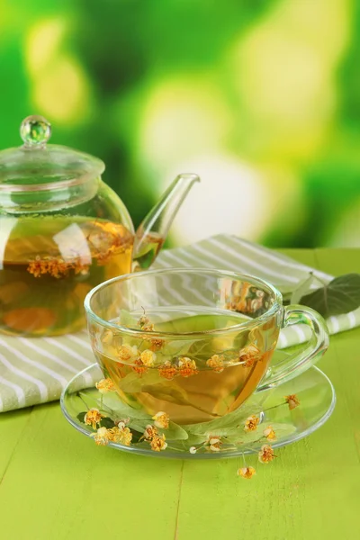 Kettle and cup of tea with linden on wooden table on nature background — Stock Photo, Image