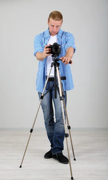 Bello fotografo con macchina fotografica su treppiede, su sfondo grigio — Foto Stock