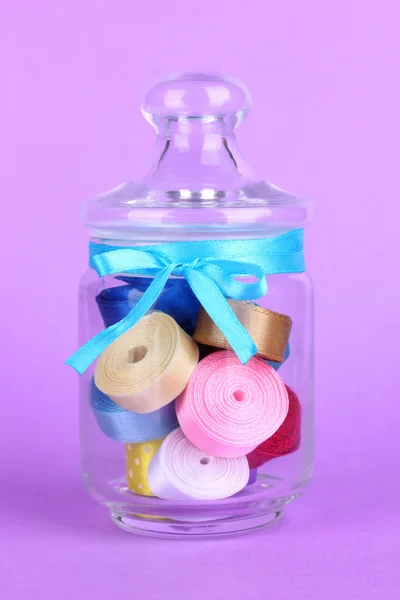 Glass jar containing various colored ribbons on lilac background — Stock Photo, Image