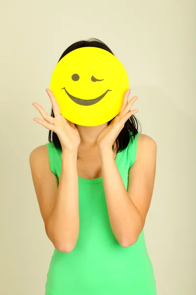 Junge Frau hält Papier mit fröhlichem Smiley auf grauem Hintergrund. — Stockfoto