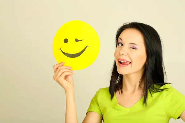 Junge Frau hält Papier mit fröhlichem Smiley auf grauem Hintergrund. — Stockfoto