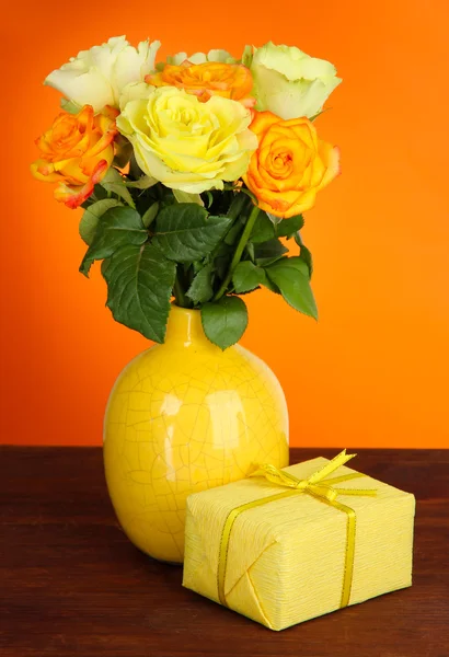 Schöner Strauß Rosen in der Vase mit Geschenk auf dem Tisch auf orangefarbenem Hintergrund — Stockfoto