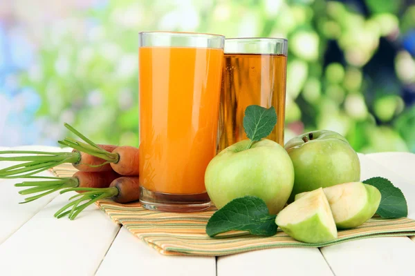 Glazen van SAP, appelen en wortelen op witte houten tafel, op groene achtergrond — Stockfoto