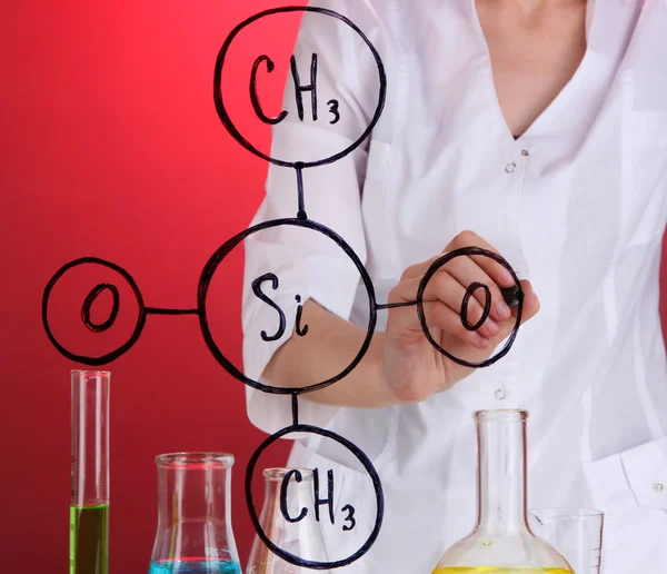 Chemist woman writing formulas on glass on red background — Stock Photo, Image