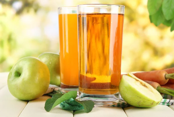 Szklanki soku, jabłka i marchew na biały drewniany stół, na zielonym tle — Zdjęcie stockowe