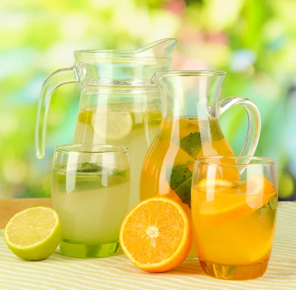 Lemoniada pomarańczowymi i cytrynowymi, dzbany i okulary na drewnianym stole na naturalne tło — Zdjęcie stockowe