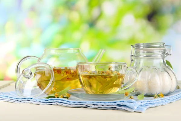 Kettle and cup of tea with linden on napkin on wooden table on nature background — Stock Photo, Image