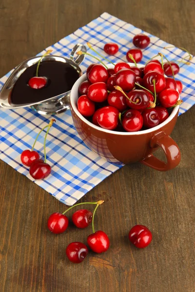 Ripe red cherry berries in cup and chocolate sauce on wooden table close-up — Stock Photo, Image
