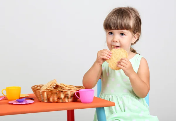 Pequena menina bonito sentado em pequena cadeira perto da mesa e comer biscoito saboroso, no fundo cinza — Fotografia de Stock