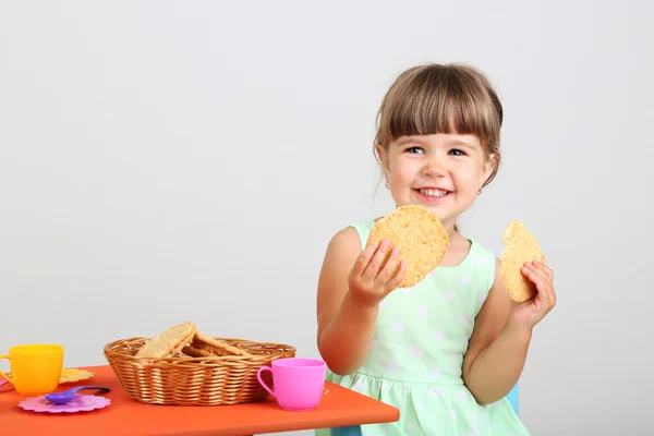 Little cute girl sitting on little chair near table and eating tasty cookie, on gray background — Stock Photo, Image